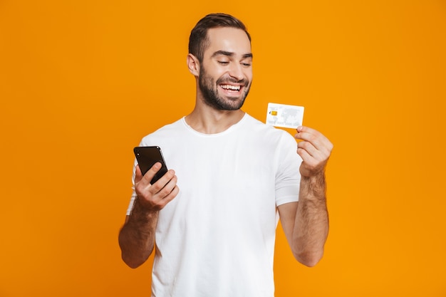 Foto de hombre alegre de 30 años en ropa casual con smartphone y tarjeta de crédito, aislado