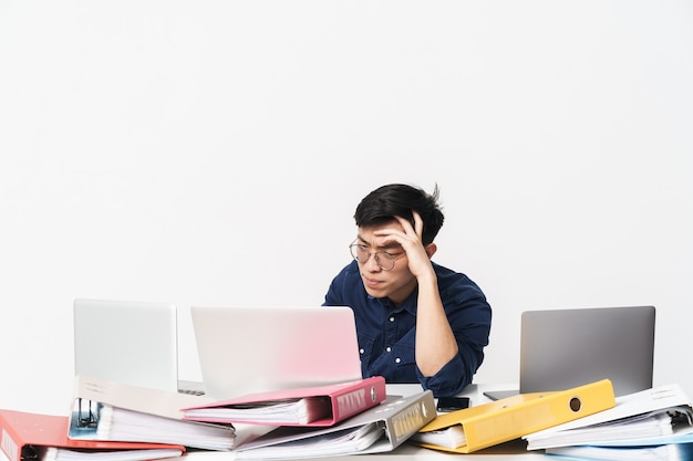Foto de hombre asiático cansado de 30 años con anteojos sentado en la mesa y usando computadoras portátiles mientras trabaja en la luminosa habitación de la oficina