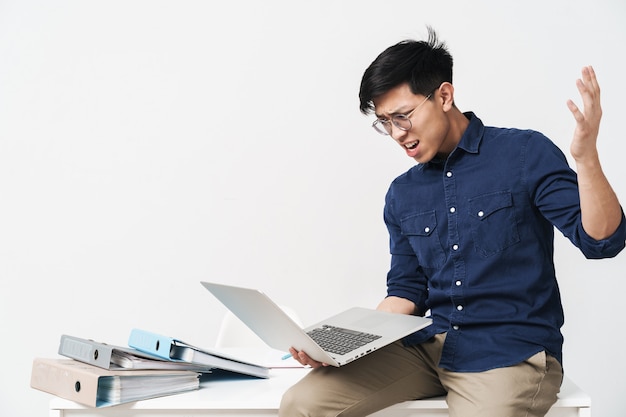 Foto de hombre asiático enojado con anteojos sentado en la mesa y trabajando con un portátil en la oficina aislada sobre la pared blanca