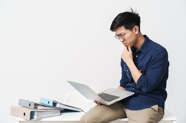 Foto de hombre asiático serio de 30 años con anteojos sentado en la mesa y escribiendo en la computadora portátil mientras trabaja en la luminosa habitación de la oficina