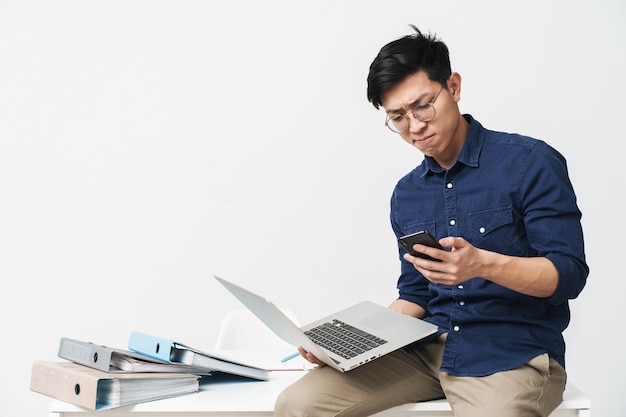 Foto de hombre asiático serio con anteojos sosteniendo un teléfono inteligente y una computadora portátil mientras trabajaba en la oficina aislada sobre la pared blanca