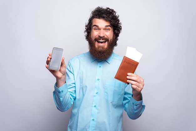 Foto de hombre asombrado que muestra la pantalla del teléfono inteligente y el pasaporte con boletos