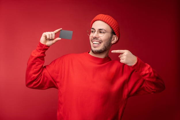 Foto de hombre atractivo con barba con gafas y ropa roja. Hombre feliz sostiene y apunta a la tarjeta de crédito, aislado sobre fondo rojo.