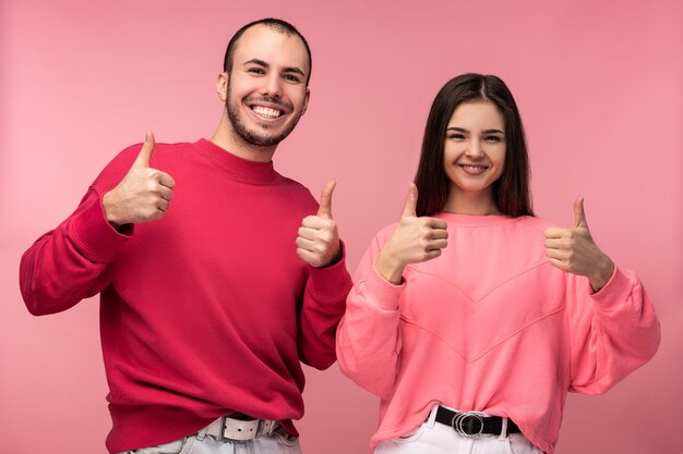 Foto foto de hombre atractivo con barba en ropa roja y mujer en rosa muestran pulgar hacia arriba y sonreír, aislado sobre fondo rosa.