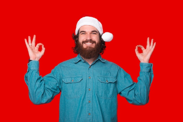 Foto de hombre barbudo alegre con sombrero de santa claus y haciendo gesto zen paz