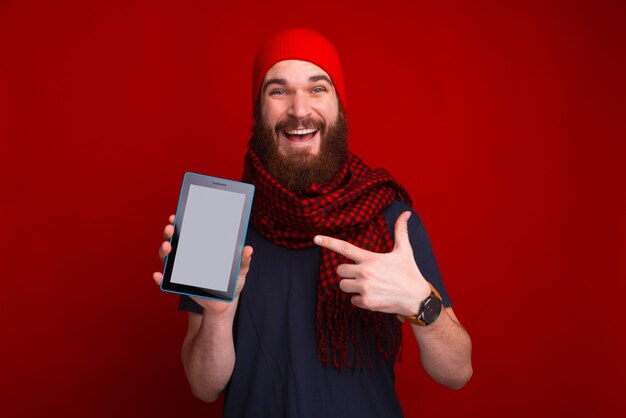 Foto de hombre barbudo feliz, con sombrero y bufanda apuntando a la pantalla de la tableta, sobre espacio rojo aislado