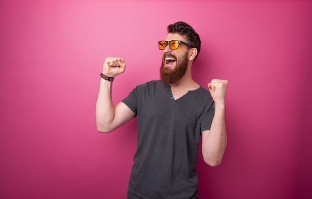 Foto de hombre barbudo ganador celebrando con manos levantadas sobre fondo rosa