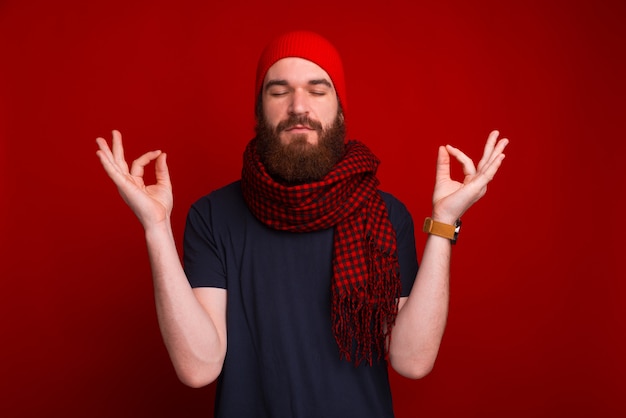 Foto de hombre barbudo, con los ojos cerrados, haciendo gesto zen, sobre espacio rojo