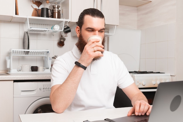 Foto de hombre bebiendo una taza de café y usando la computadora portátil en la cocina