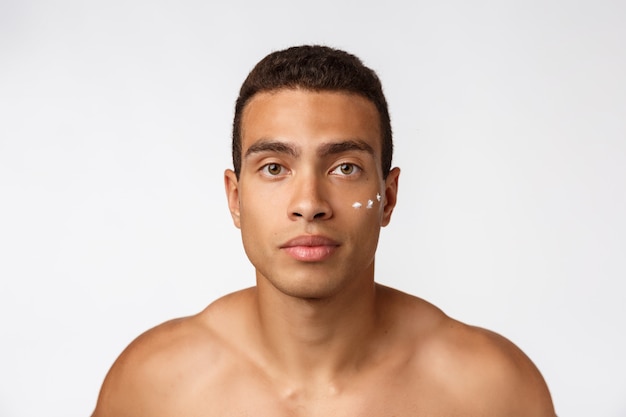 Foto de hombre sin camisa sonriendo y aplicando crema para la cara aislado en blanco