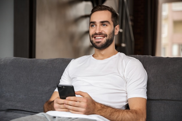 Foto de hombre caucásico de 30 años vistiendo camiseta casual sosteniendo y usando el teléfono inteligente mientras está sentado en el sofá en la sala de estar