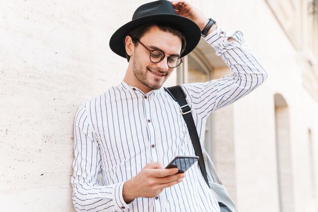 Foto de hombre caucásico alegre con anteojos y sombrero negro escribiendo en el teléfono celular y sonriendo mientras se inclina en la pared en las calles de la ciudad