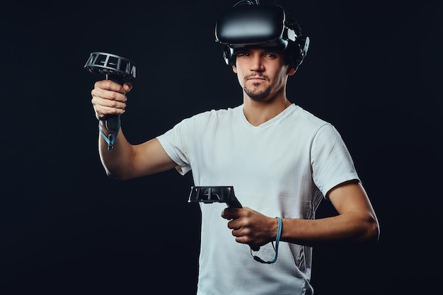 Foto de un hombre caucásico con cerdas vestido con camisa blanca usando gafas 3d con joysticks. Aislado sobre fondo oscuro.