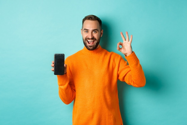 Foto de hombre caucásico que muestra la pantalla del móvil y el signo bien, aprueba la tienda en línea, la aplicación de teléfono inteligente, de pie satisfecho sobre el fondo azul claro.
