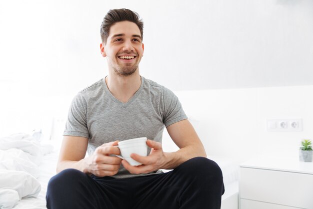 Foto foto del hombre complacido en la mañana sonriendo mientras estaba sentado en el dormitorio y bebiendo té o café