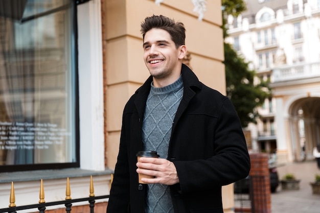 Foto de hombre elegante de 30 años con chaqueta sosteniendo una taza de papel de café para llevar mientras camina por las calles de la ciudad