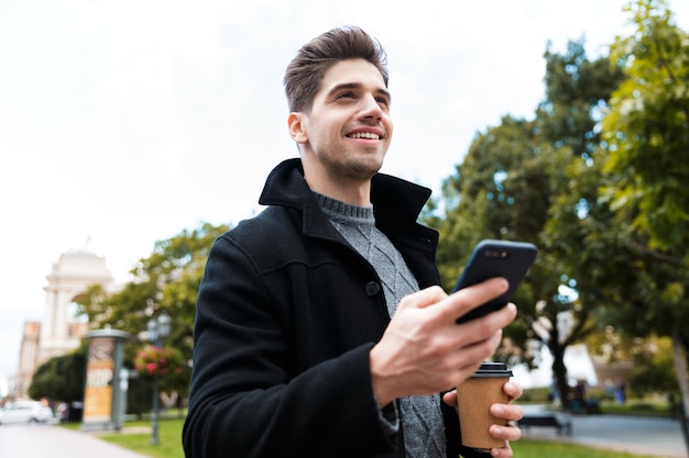 Foto de hombre elegante de 30 años con chaqueta sosteniendo teléfono inteligente y bebiendo café para llevar mientras camina por el parque de la ciudad