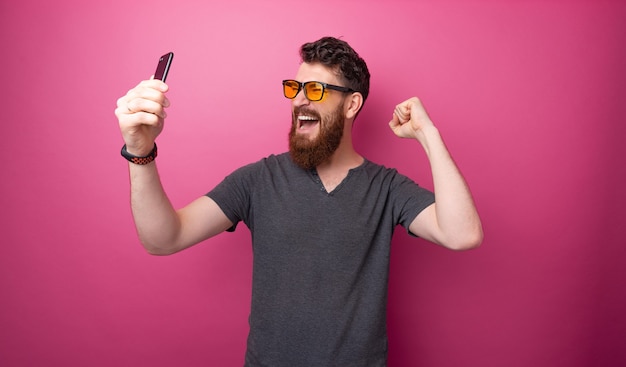 Foto de hombre emocionado con barba y gafas de sol tylish, sosteniendo el teléfono móvil mientras celebra con las manos levantadas