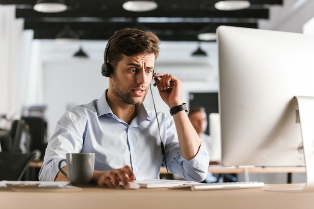 Foto foto de hombre estresado de 30 años con ropa de oficina y auriculares, sentado junto a la computadora y trabajando en el centro de llamadas