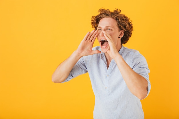 Foto de hombre europeo feliz gritando o llamando mientras sostiene las manos en la cara, aislado sobre fondo amarillo