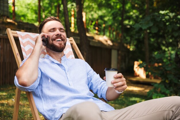 Foto de hombre europeo feliz riendo y sentado en un sillón con café para llevar durante el descanso en el parque