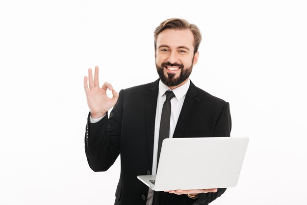 Foto de hombre exitoso en traje negro sonriendo y mostrando un signo OK mientras sostiene el cuaderno, aislado sobre la pared blanca