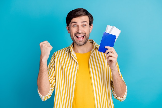 Foto de hombre extasiado ganar en el extranjero viaje lotería de viaje mantenga los billetes levantar puños usar ropa blanca aislada sobre fondo de color azul