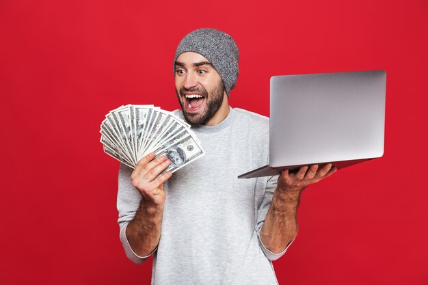 Foto de hombre feliz de 30 años en ropa casual con dinero en efectivo y portátil plateado aislado