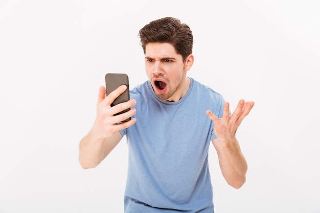 Foto de un hombre frustrado de 30 años gritando por teléfono celular cuando recibe noticias molestas o mensajes desagradables, aislado sobre una pared blanca