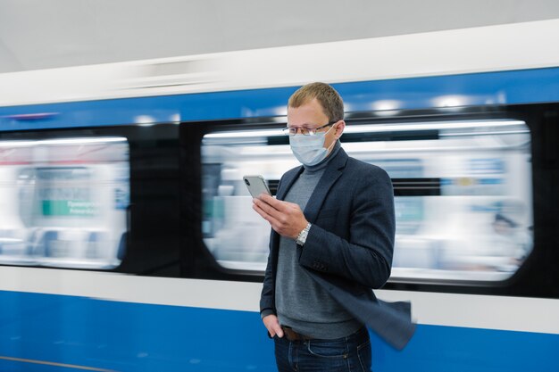 Foto del hombre con gafas y máscara médica protectora, usa un teléfono moderno, viaja en transporte público, se para contra el tren en movimiento en la plataforma, obtiene información en línea sobre el nuevo virus Covid-19