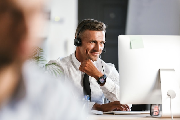 Foto de hombre guapo de 30 años con ropa de oficina y auriculares, trabajando en la computadora en el centro de llamadas