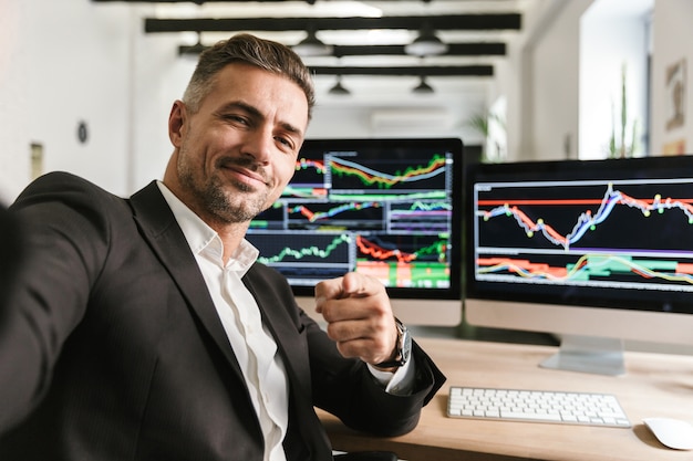 Foto de hombre guapo de 30 años con traje tomando selfie mientras trabaja en la oficina en la computadora con gráficos y tablas en la pantalla