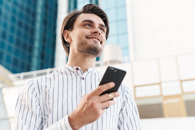 Foto de hombre guapo alegre con camisa a rayas escribiendo en el teléfono celular y sonriendo mientras camina por las calles de la ciudad