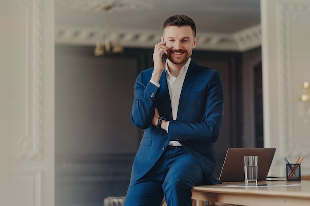 Foto de hombre guapo con barba tiene conversación telefónica hace llamada celular satisfecha con buenas noticias posa en el escritorio con computadora portátil moderna vestida con traje formal azul. Conversación móvil