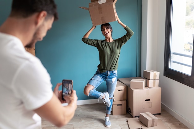 Foto de un hombre guapo tomando una foto con un teléfono inteligente a su divertida esposa mientras bromea con las cajas de cartón en movimiento en la nueva casa.