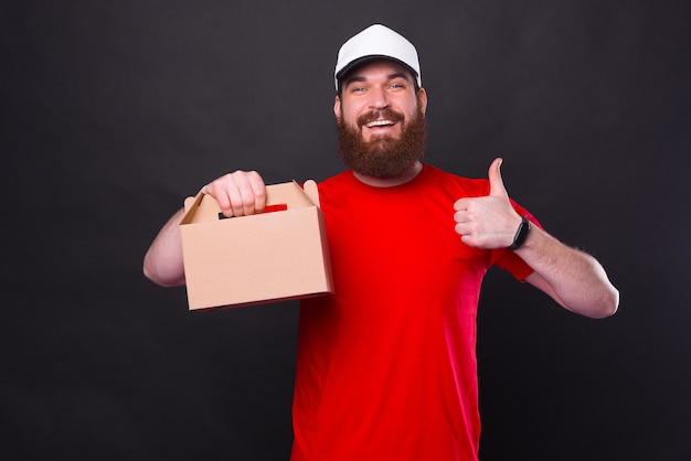 Foto de hombre hipster barbudo joven alegre en camiseta roja mostrando el pulgar hacia arriba y lonchera