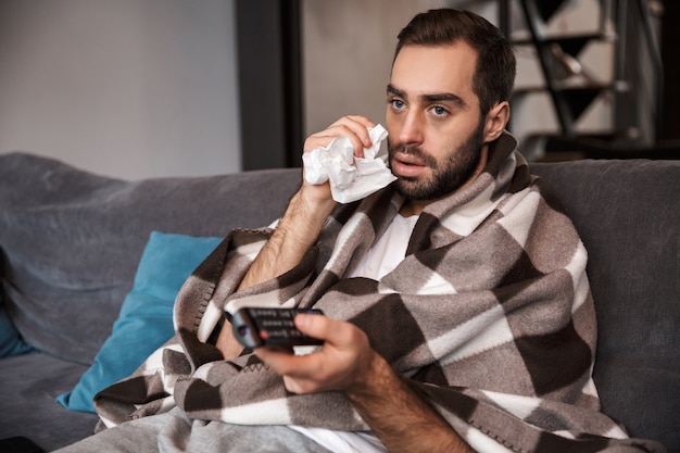 Foto de hombre insatisfecho de 30 años que tiene temperatura y está enfermo mientras está sentado envuelto en una manta en el sofá en casa