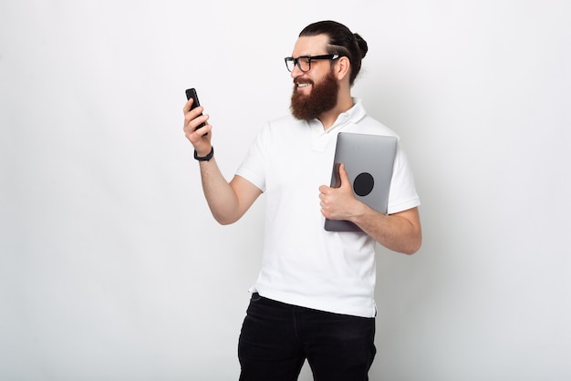 Foto de hombre joven con barba en camiseta blanca con smartphone y portátil