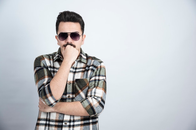 Foto de hombre joven con gafas negras de pie sobre una pared blanca.