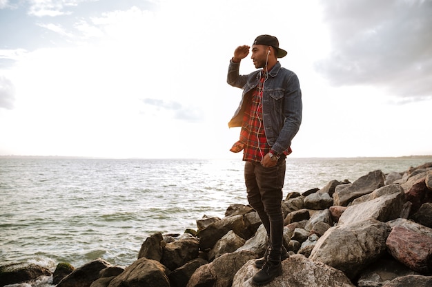 Foto de hombre joven con gorra caminando por la playa mientras escucha música con auriculares.