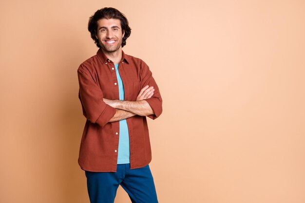 Foto de hombre joven mirada segura de sí mismo con las manos cruzadas de la cámara confía en llevar camiseta marrón pantalones azules aislados de fondo de color beige