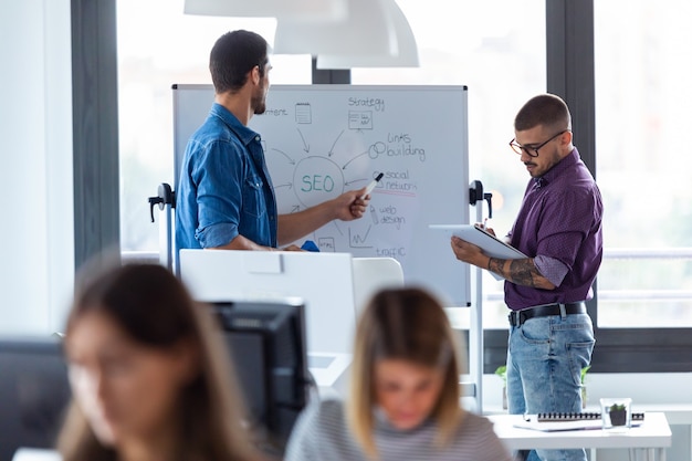 Foto de hombre joven de negocios apuntando en la pizarra mientras habla con su colega del nuevo proyecto en la oficina de inicio moderna.