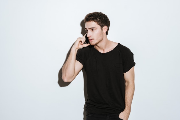 Foto de hombre joven vestido con camiseta negra posando sobre fondo blanco y hablando por teléfono. Mirando a un lado.