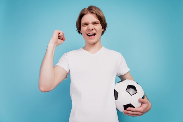 Foto de hombre loco positivo sosteniendo un balón de fútbol levantar el puño aislado fondo azul.