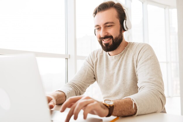 Foto de hombre maduro atractivo en ropa casual escribiendo mensajes de texto o trabajando en la computadora portátil, mientras escucha música a través de auriculares