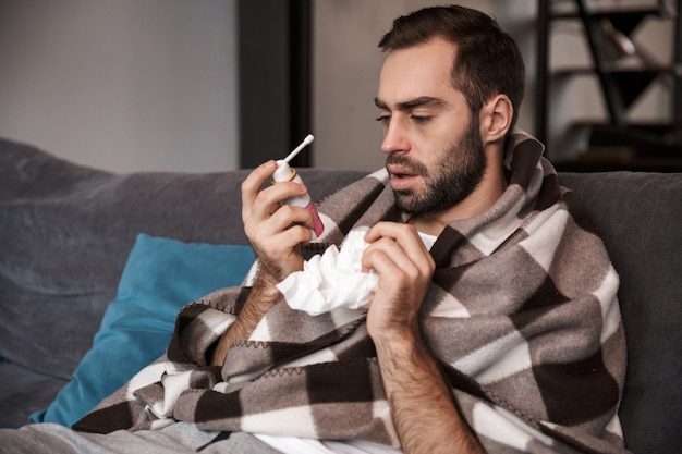 Foto de hombre molesto de 30 años envuelto en una manta que tiene temperatura y está enfermo mientras está sentado en el sofá en el apartamento