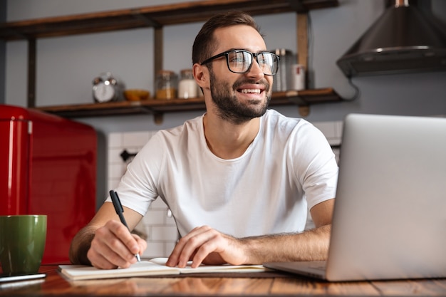 Foto de hombre morena de 30 años con anteojos anotando notas mientras usa el portátil plateado en la mesa de la cocina