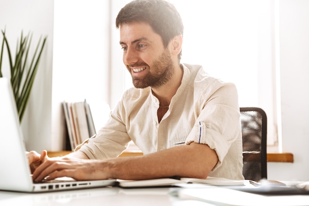 Foto de hombre de negocios alegre de 30 años con camisa blanca trabajando con una computadora portátil y documentos en papel, mientras está sentado en la oficina brillante