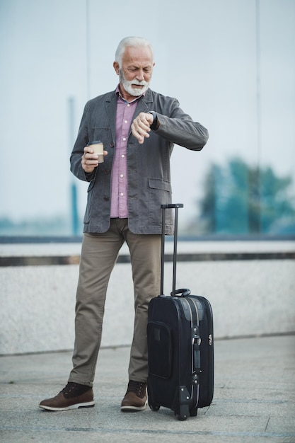 Una foto de un hombre de negocios de alto rango parado con su equipaje y mirando un reloj de mano fuera del aeropuerto.