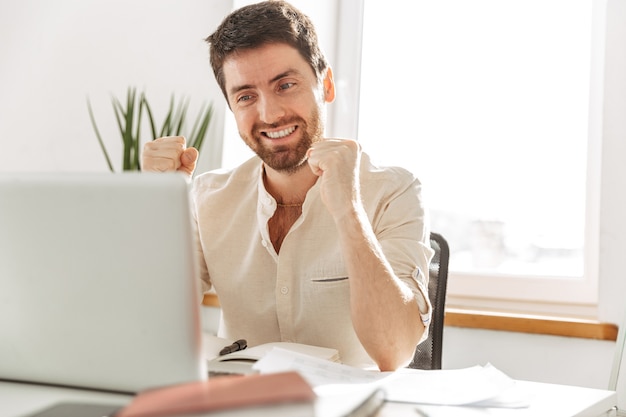 Foto de hombre de negocios feliz de 30 años vistiendo camisa blanca con portátil, mientras trabaja en la oficina